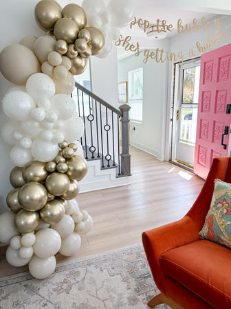 Home entryway decorated with a white and gold balloon garland around a staircase, bright pink front door, glitter birthday banner overhead, and an orange accent chair with patterned pillow.