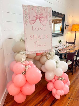 Pastel pink and white balloon garland with baby's-breath accents framing a floral sign reading "LOVE SHACK Fiancé" beside a rustic wooden-floor dining table set for an indoor engagement party