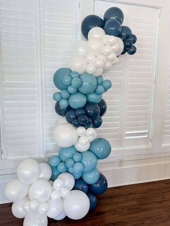 Playful indoor balloon garland in clustered shades of white, sky blue and navy forming a tall curving column against white plantation shutters over a hardwood floor.