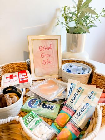 Woven guest amenity basket on a white table with a vase of greenery and a pink framed sign reading “Refresh Renew Relax,” filled with hydration powder sticks, gum, mints, first-aid kit, hair ties and under-eye patches.