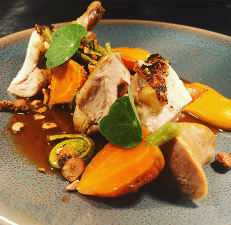 Fine-dining roasted chicken with crispy skin, glazed carrots, fiddlehead ferns, toasted hazelnuts and brown jus, garnished with green nasturtium leaves on a speckled blue plate