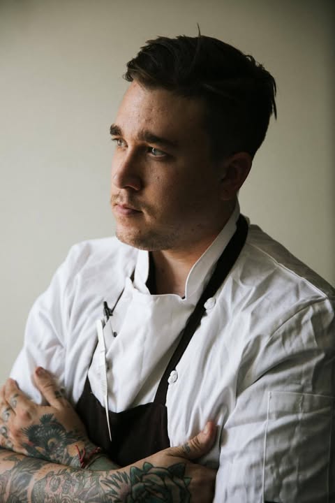 Close-up portrait of a tattooed chef wearing a white jacket and dark apron with arms crossed, looking thoughtful