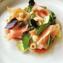 Gourmet Italian-style starter: thin prosciutto slices draped over ripe tomato rounds with fresh basil and mint leaves, toasted hazelnuts and olive oil on a white plate.