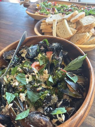 Hearty wooden bowl of steamed mussels tossed with fresh basil, herbs and shredded seafood, served alongside sliced crusty bread and a leafy salad in wooden bowls on a rustic table