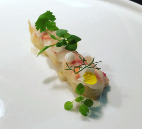 Minimalist fine-dining raw seafood crudo on a white plate, topped with microgreens, citrus pearls and a small yellow edible flower.