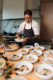 Professional chef plating creamy mushroom risotto into white bowls on a wooden counter in a modern kitchen, bowls lined up and ready for service