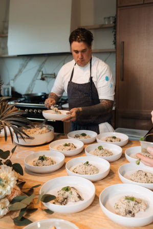 Professional chef plating creamy mushroom risotto into white bowls on a wooden counter in a modern kitchen, bowls lined up and ready for service