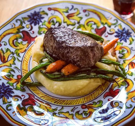 Seared steak atop creamy mashed potatoes with glazed baby carrots and green beans served on a vibrant hand-painted ceramic plate
