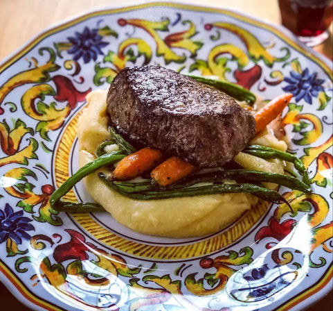 Seared steak atop creamy mashed potatoes with glazed baby carrots and green beans served on a vibrant hand-painted ceramic plate