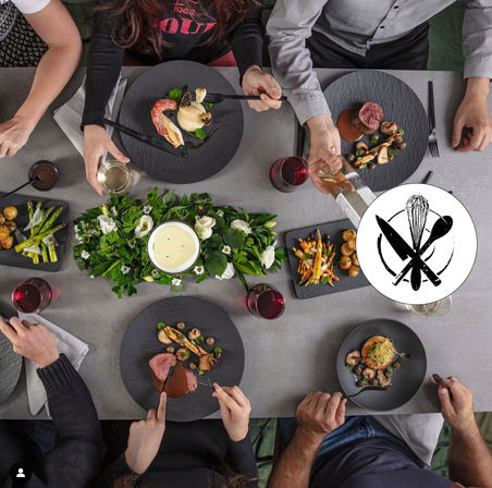Overhead view of a modern dinner party: people sharing restaurant-style plated gourmet dishes on slate plates — seared steak with mushrooms, prawn with puree, roasted vegetables, asparagus and baby potatoes — red and white wine glasses and a green floral candle centerpiece on a gray table.