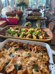 Cheesy baked tortellini casserole topped with fresh basil in a white dish, with wooden platters of marinated vegetables, olives and greens, plus a fruit bowl and bananas on a long sunlit dining table in a buffet-style spread.