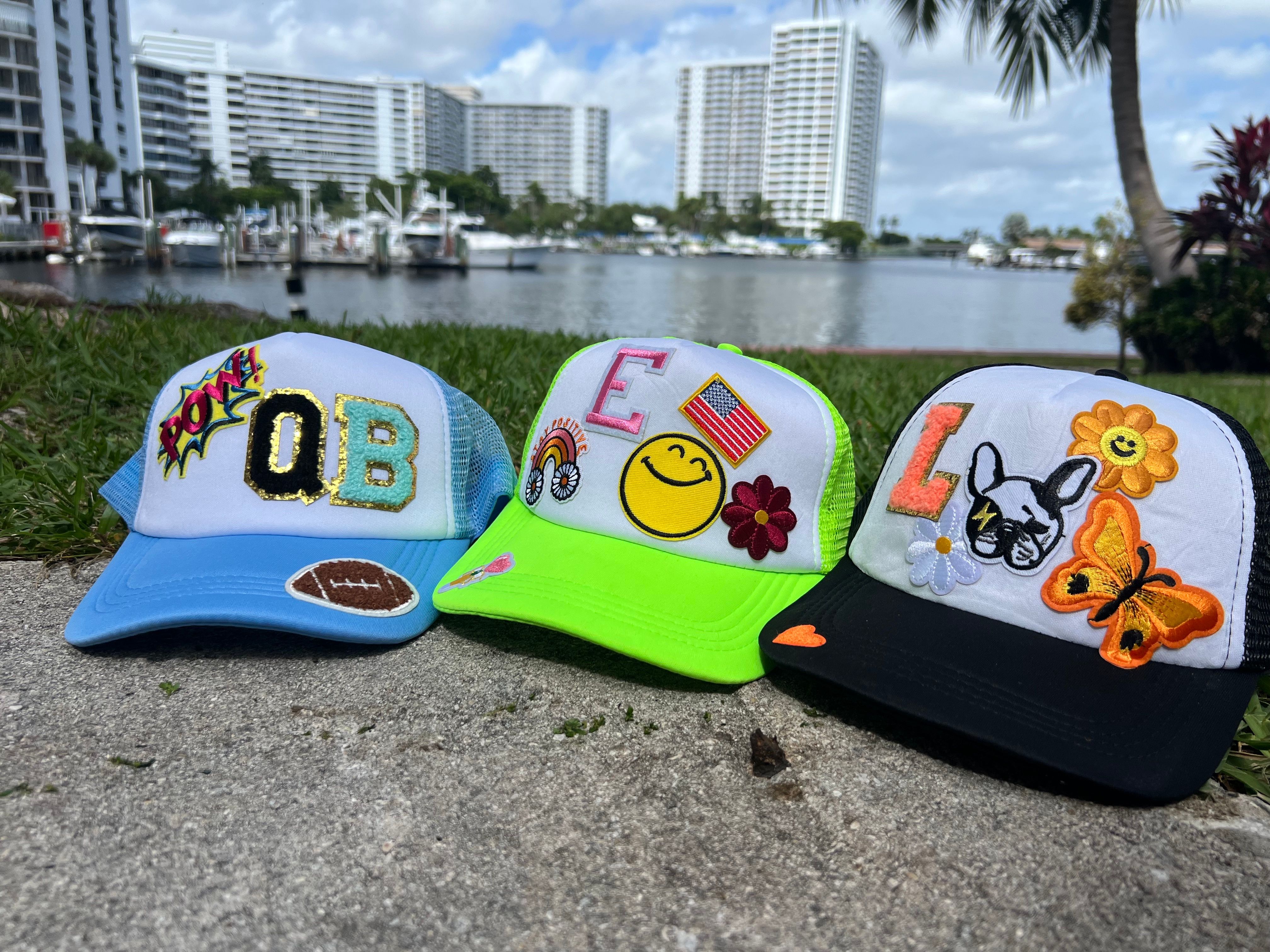 Three colorful custom trucker hats with patches — blue 'QB' football cap, neon green smiley‑flag cap, and black butterfly cap — displayed on a waterfront path with boats and high‑rise condos in the background.