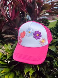 Bright pink and white trucker hat with embroidered colorful daisies and a red heart, photographed outdoors resting on tropical green foliage