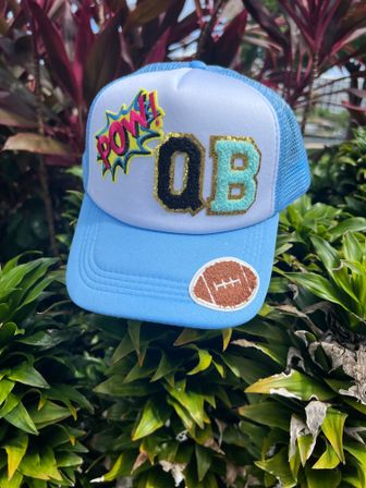Playful light-blue trucker hat with bright 'POW!' comic patch, gold-trimmed 'QB' chenille letters and a small football patch resting on lush tropical garden foliage