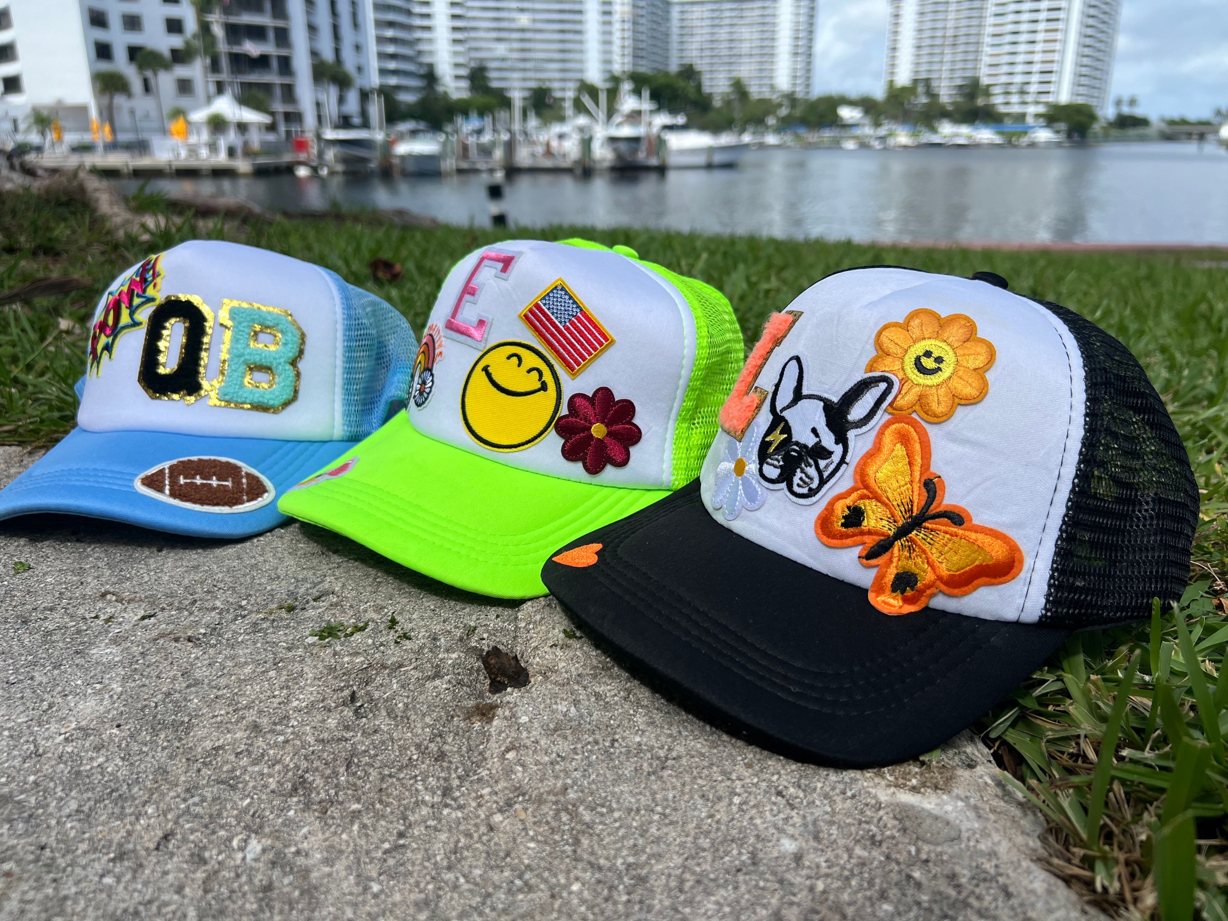 Three colorful trucker hats with embroidered patches — blue 'QB' with a football, neon green smiley with an American flag, and black-and-white cap with a butterfly and dog — lined up on a concrete waterfront path by a marina and high-rise condos.