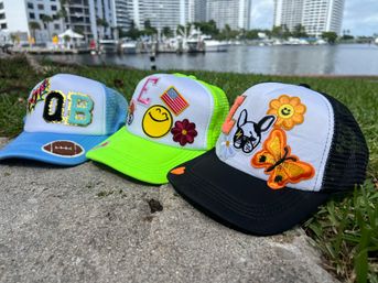 Three colorful trucker hats with playful embroidered patches — blue 'QB' with a football, neon green with smiley and flag, and black-and-white with butterfly and dog — lined up on a waterfront sidewalk by a marina with boats and high-rise condos in the background.