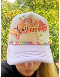 Close-up of a person wearing a pastel pink trucker hat decorated with patches reading "Spread Good Vibes", a butterfly, daisy and smiling heart, plus a braided rainbow trim, photographed outdoors against green foliage.
