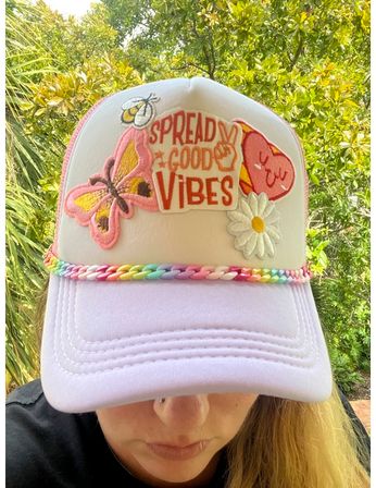Close-up of a person wearing a pastel pink trucker hat decorated with patches reading "Spread Good Vibes", a butterfly, daisy and smiling heart, plus a braided rainbow trim, photographed outdoors against green foliage.