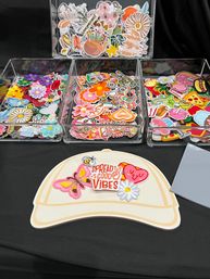 Colorful embroidered iron-on patches—flowers, butterflies, hearts, pizza and more—stacked in clear acrylic trays on a black table with a hat-shaped display reading 'Spread Good Vibes' at a craft-market style vendor table