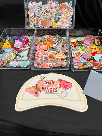 Colorful embroidered iron-on patches—flowers, butterflies, hearts, pizza and more—stacked in clear acrylic trays on a black table with a hat-shaped display reading 'Spread Good Vibes' at a craft-market style vendor table