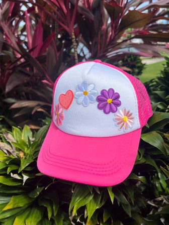 Pink mesh trucker hat with white front panel embroidered with colorful daisies and a red heart, photographed resting on green tropical plants in an outdoor garden.