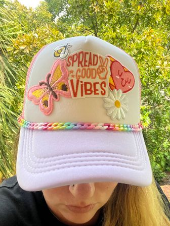 Close-up of a pastel trucker hat with a lavender brim and white front, featuring embroidered patches reading "Spread Good Vibes" with a pink butterfly, daisy and heart, rainbow plastic chain trim, worn outdoors against green leafy background.