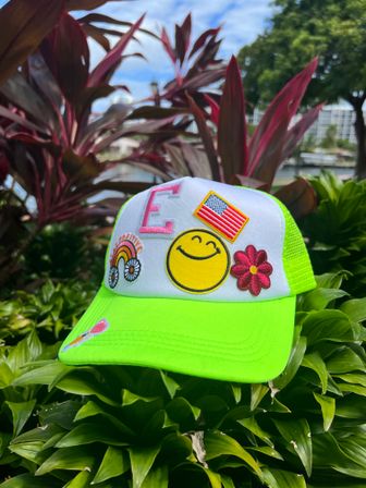 Neon green trucker hat with white front panel decorated with patches — yellow smiley face, American flag, pink letter E, rainbow daisies and a pink flower — resting on tropical green foliage with red-leaf plants and a blurred waterfront in the background.