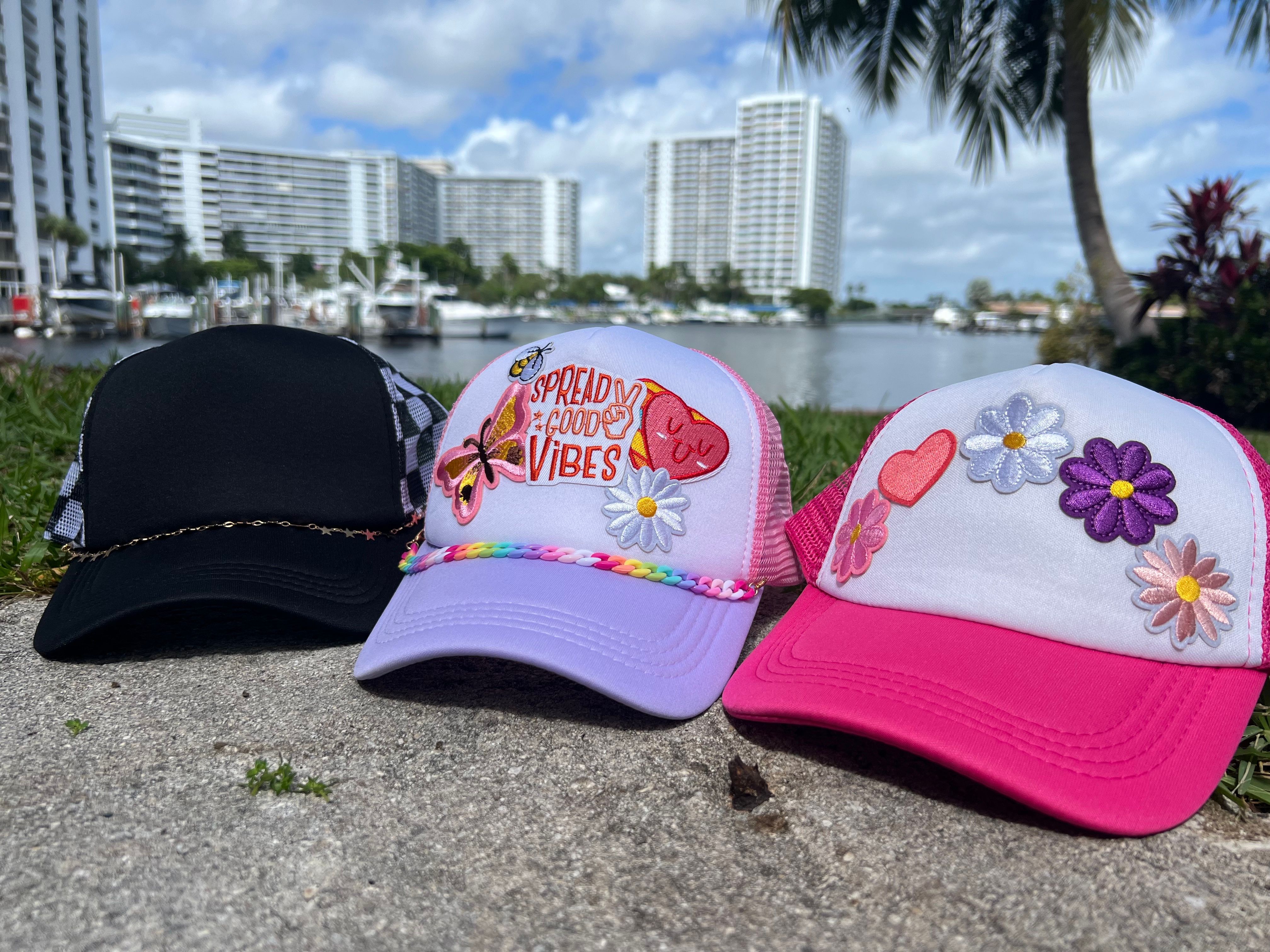 Three embroidered trucker hats — black, lavender with a “Spread Good Vibes” patch, and hot pink with flower patches — photographed on a marina waterfront with palm trees and high-rise condos.