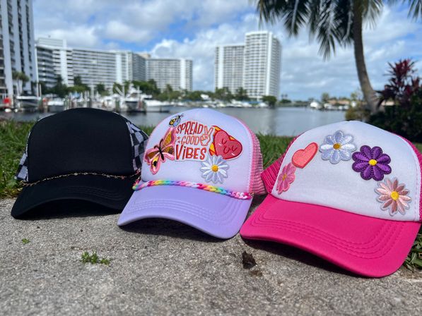 Three colorful trucker hats—black, pastel purple with a "Spread Good Vibes" patch, and bright pink with floral patches—resting on a waterfront concrete ledge with yachts and high-rise condos in the background.