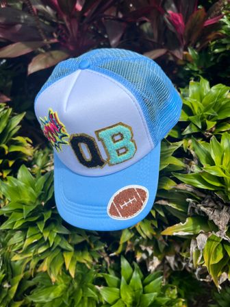 Light blue mesh trucker hat with embroidered QB letters, comic-style burst patch and a football patch, photographed on lush tropical green foliage