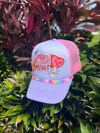 Lavender and pink trucker hat with embroidered "Spread Good Vibes" text, daisy and heart patches and a rainbow chain on the brim, photographed on lush tropical foliage in a garden
