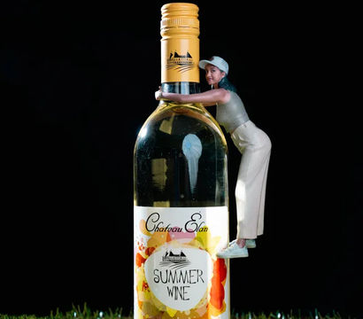 Miniature woman in a cap hugging an oversized bottle of white wine labeled Summer Wine, standing on green grass against a black background