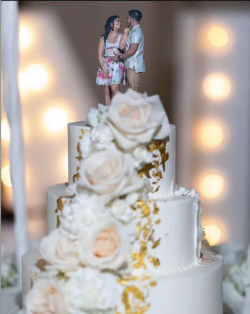 Three-tier white wedding cake with cascading blush roses and gold leaf accents, topped by a smiling couple figurine cake topper against warm bokeh lights