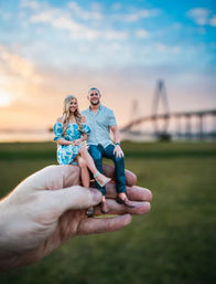 Hand holding tiny custom 3D-printed figurines of a smiling couple sitting on a palm, grassy waterfront park and a silhouetted bridge at sunset