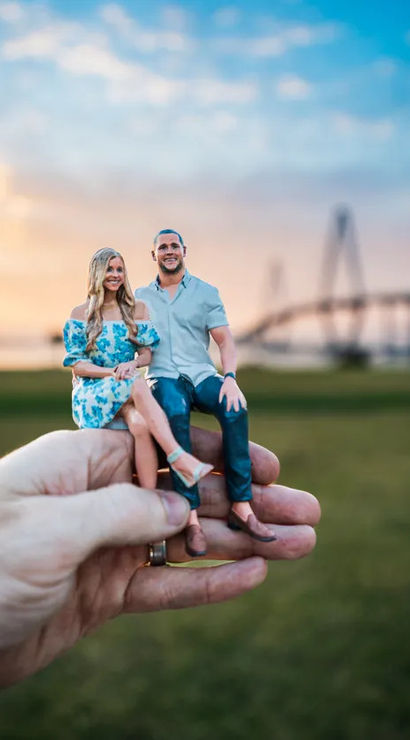 Hand holding tiny custom 3D-printed figurines of a smiling couple sitting on a palm, grassy waterfront park and a silhouetted bridge at sunset