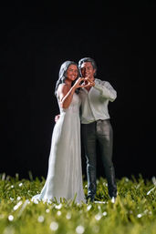 Miniature wedding couple figurine on grass, bride in a white gown and groom in shirt and trousers forming a heart with their hands — romantic cake topper style shot
