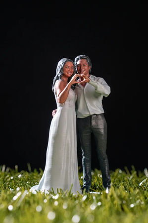 Miniature wedding couple figurine on grass, bride in a white gown and groom in shirt and trousers forming a heart with their hands — romantic cake topper style shot