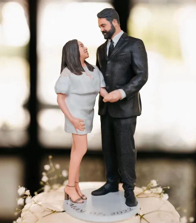 Wedding cake topper showing a miniature bearded man in a black suit holding hands with a woman in a short white dress, standing on a cake decorated with baby's-breath flowers and a blurred indoor backdrop.