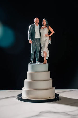 Realistic couple figurine cake topper on a modern four-tier white fondant wedding cake, woman in a slip dress and man in a suit against a dramatic black background