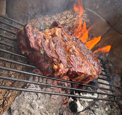 Close-up of a juicy, charred steak sizzling on a grill grate over glowing embers and orange wood flames in an outdoor wood‑fire barbecue.