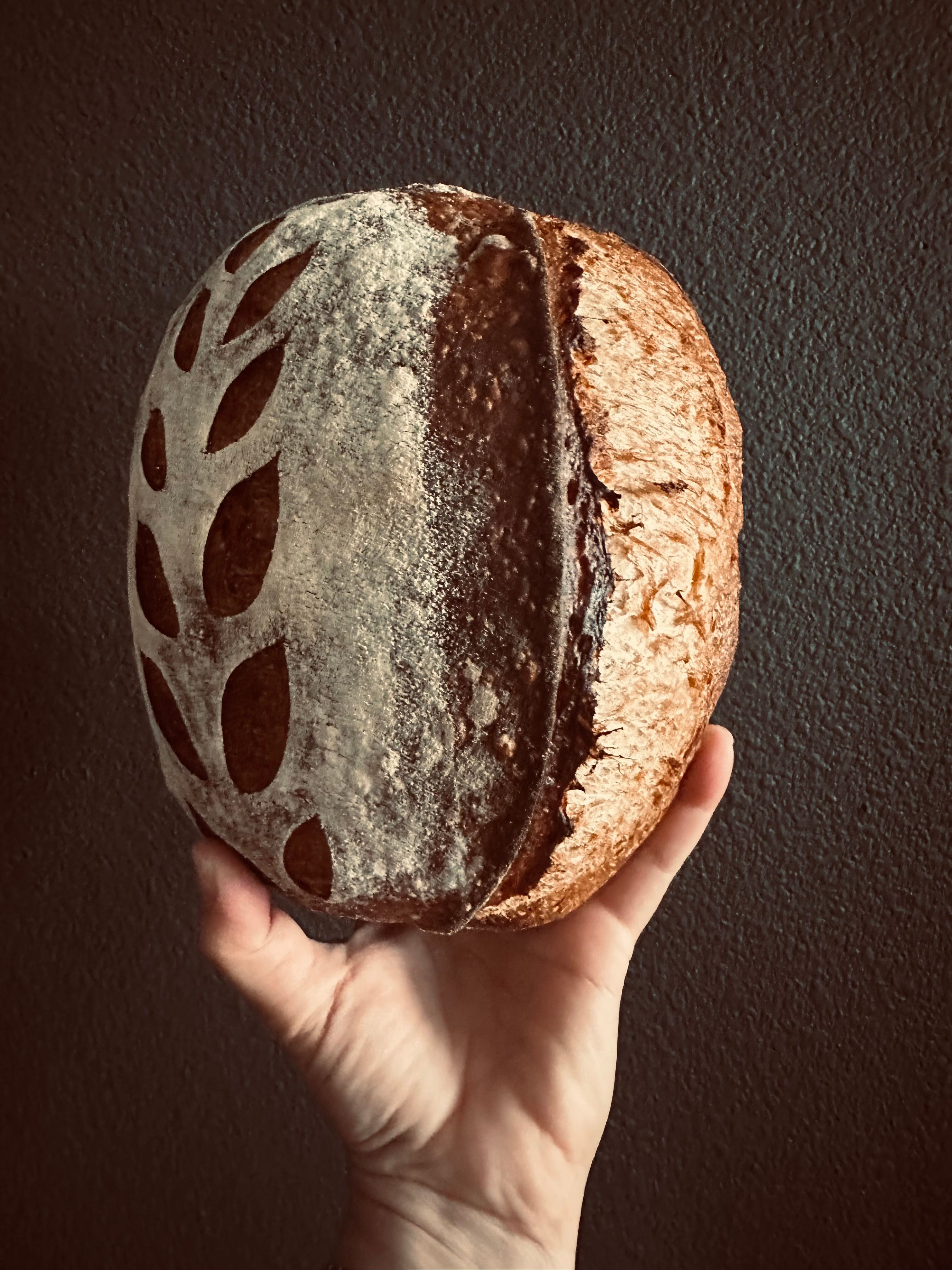 Hand holding an artisan sourdough loaf with decorative leaf scoring, flour-dusted dark crust and golden-brown side against a textured dark wall — rustic homemade bread.