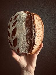 Hand holding an artisan sourdough loaf with decorative leaf scoring, flour-dusted dark crust and golden-brown side against a textured dark wall — rustic homemade bread.
