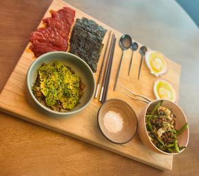 Top-down view of an Asian-inspired wooden serving board with an avocado rice bowl topped with sesame and herbs, tuna sashimi slices, nori sheets, halved soft‑boiled eggs, a small salt dish, grilled asparagus salad and chopsticks.