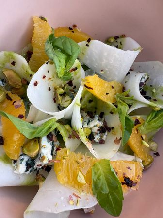Close-up citrus salad with shaved white root vegetable ribbons, juicy orange segments, crumbled blue cheese, chopped pistachios, fresh basil leaves and red pepper flakes in a pink bowl