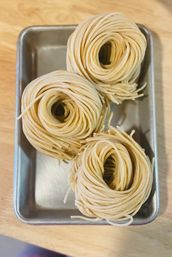 Three nests of fresh uncooked egg pasta neatly coiled in a metal tray on a wooden kitchen countertop.