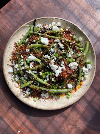 Charred blistered green beans with crunchy red chili crumbs, crumbled feta and sliced scallions on a rustic ceramic plate atop a wooden table