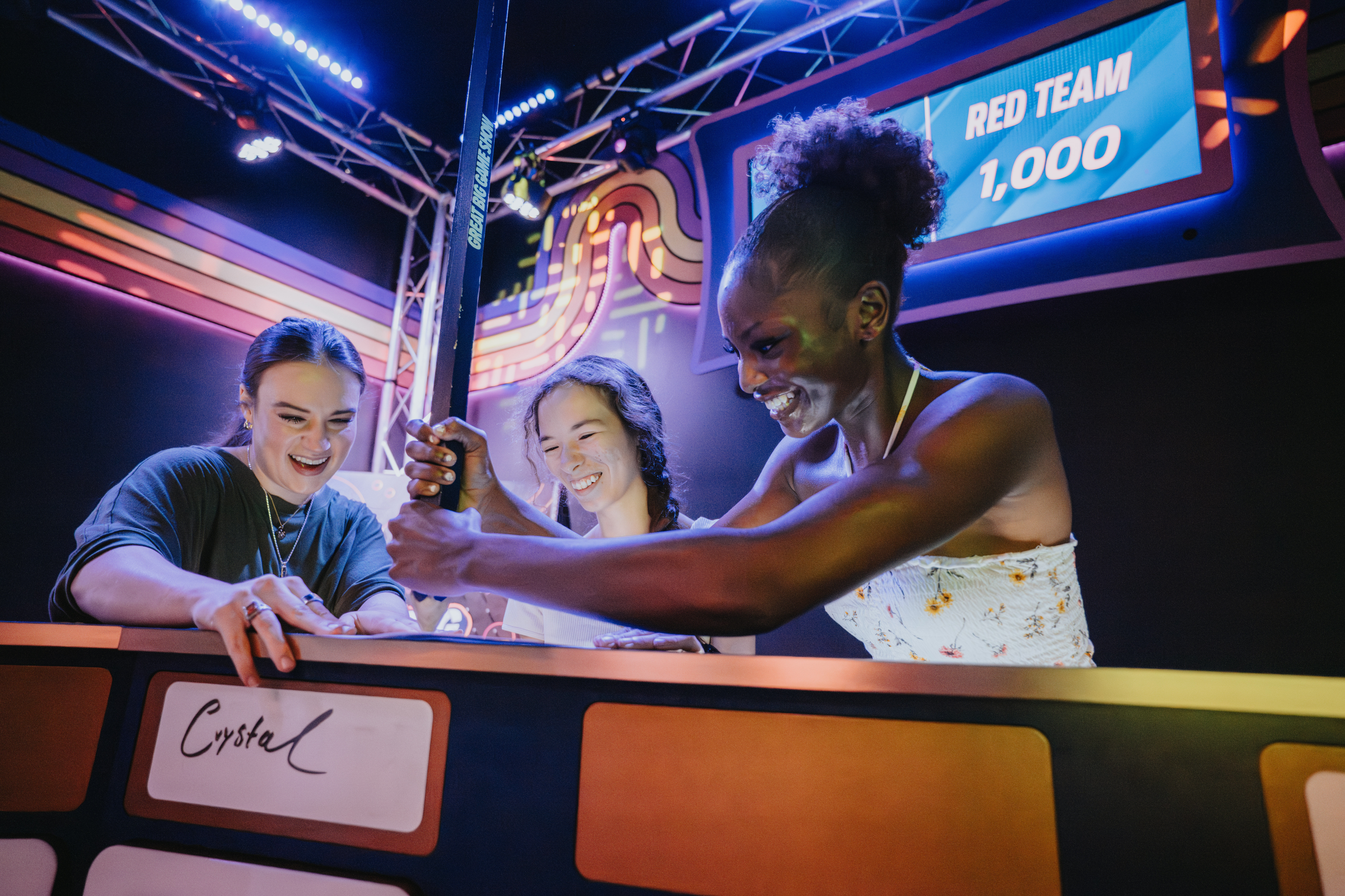 Three women laughing as they compete in a neon-lit arcade-style game show, one pulling a lever while a scoreboard reads “Red Team 1,000”.