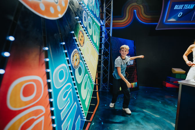 Child playing a colorful, lighted arcade-style game wall inside an indoor family entertainment center, mid-throw with a game-show scoreboard in the background.