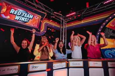 Five women cheering and raising their arms on a colorful game-show stage with studio lights and falling confetti, a bright marquee above reads "RED TEAM WINS!" over a contestants’ podium.