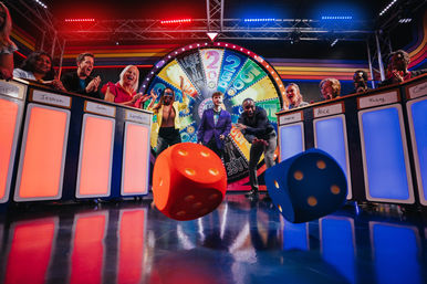 Excited contestants on a colorful TV game show stage with a spinning prize wheel and giant red and blue dice rolling across the glossy studio floor.